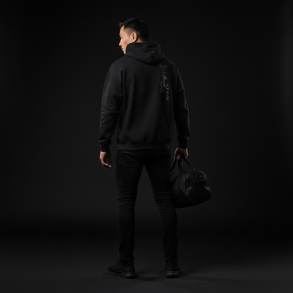 Person wearing a black hoodie and pants holding a black bag against a dark background, black magic hoodie