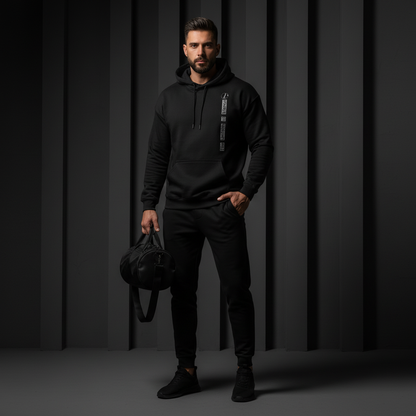 Man wearing a black hoodie and pants holding a black bag against a dark background, the uniform of silence