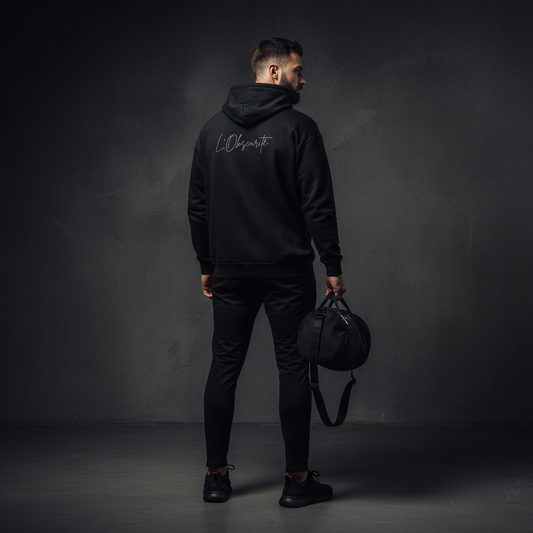 Man wearing a black hoodie, holding a black duffel bag against a dark background. Urban streetwear.